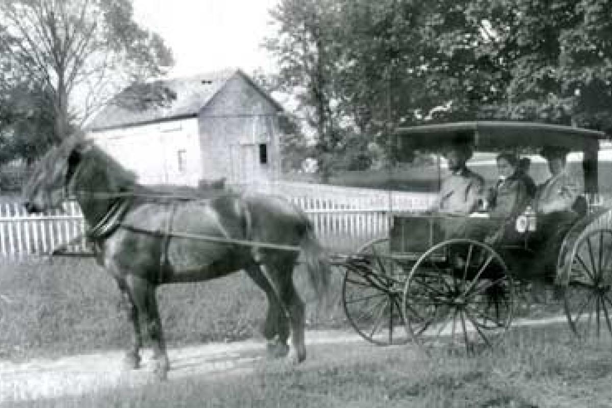 Circa 1905 photo of Alden Reynolds driving his carriage with son Hayden next to him—wife Mary Louise Whiting just behind—last person is unidentified—location is Bushnell Street with Dickinson Barn in center background—current Teal Lane area and North Cove may be seen in background.